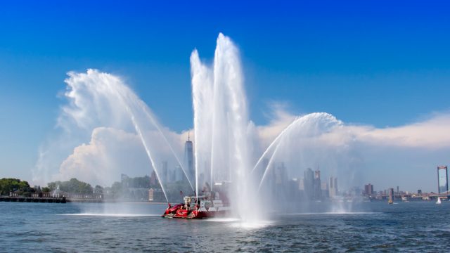 NYFD fireboat sprays a watery salute to the Brooklyn E-Prix race. In back, Governors Island and Manhattan&#039;s Wall Street, with Freedom Tower standing tall between the sprays. (Photo: Bill Howard)